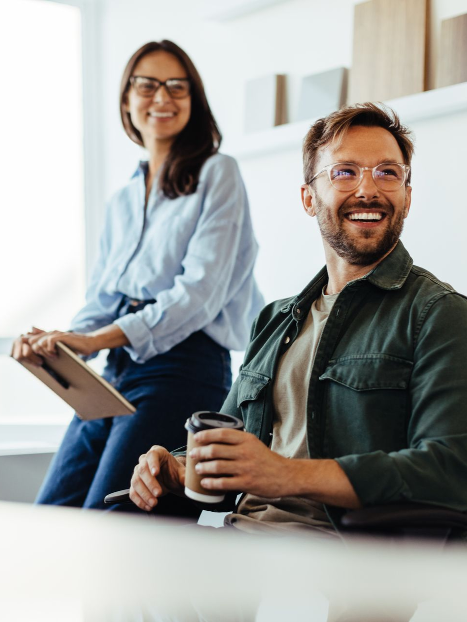Woman with glasses holds notepad behind smiling man with coffee.