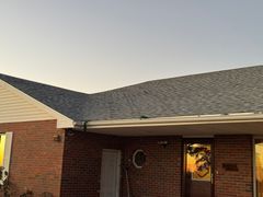 Brick house with dark gray shingle roof and golden-lit windows during sunset.