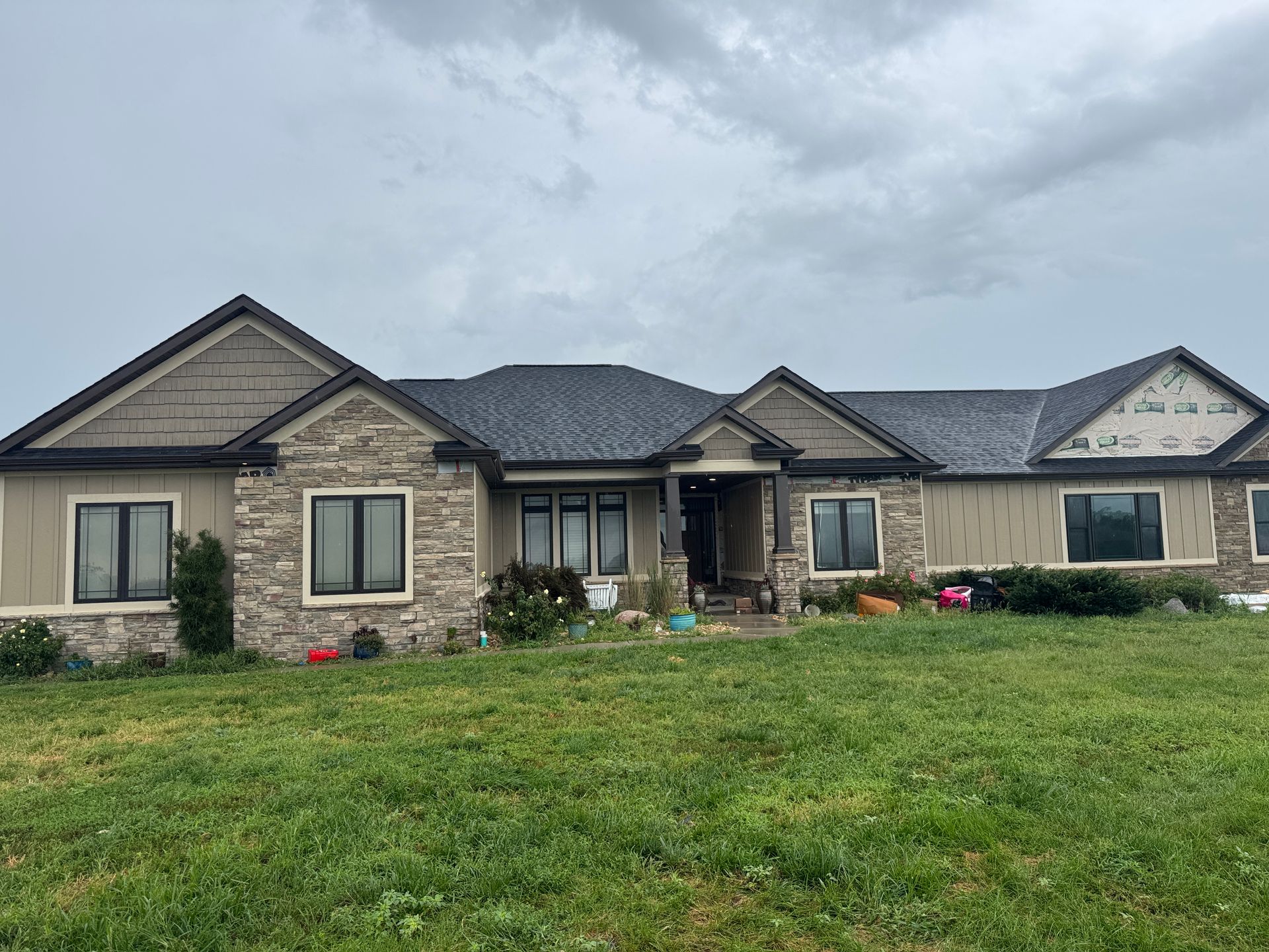 Ranch-style house with stone and tan siding, black windows, and a dark roof on a cloudy day.