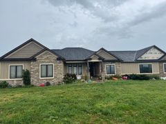 Ranch-style house with stone and tan siding, black windows, and a dark roof on a cloudy day.