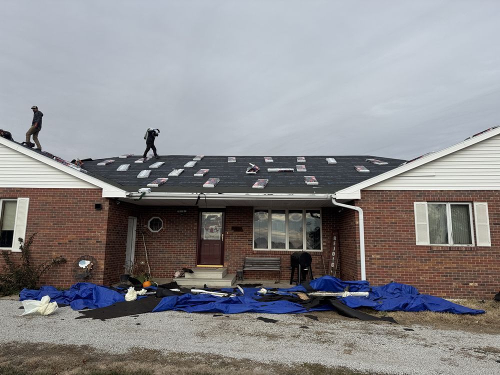 Two roofers work on a damaged brick house with blue tarp on the ground. Gray sky.