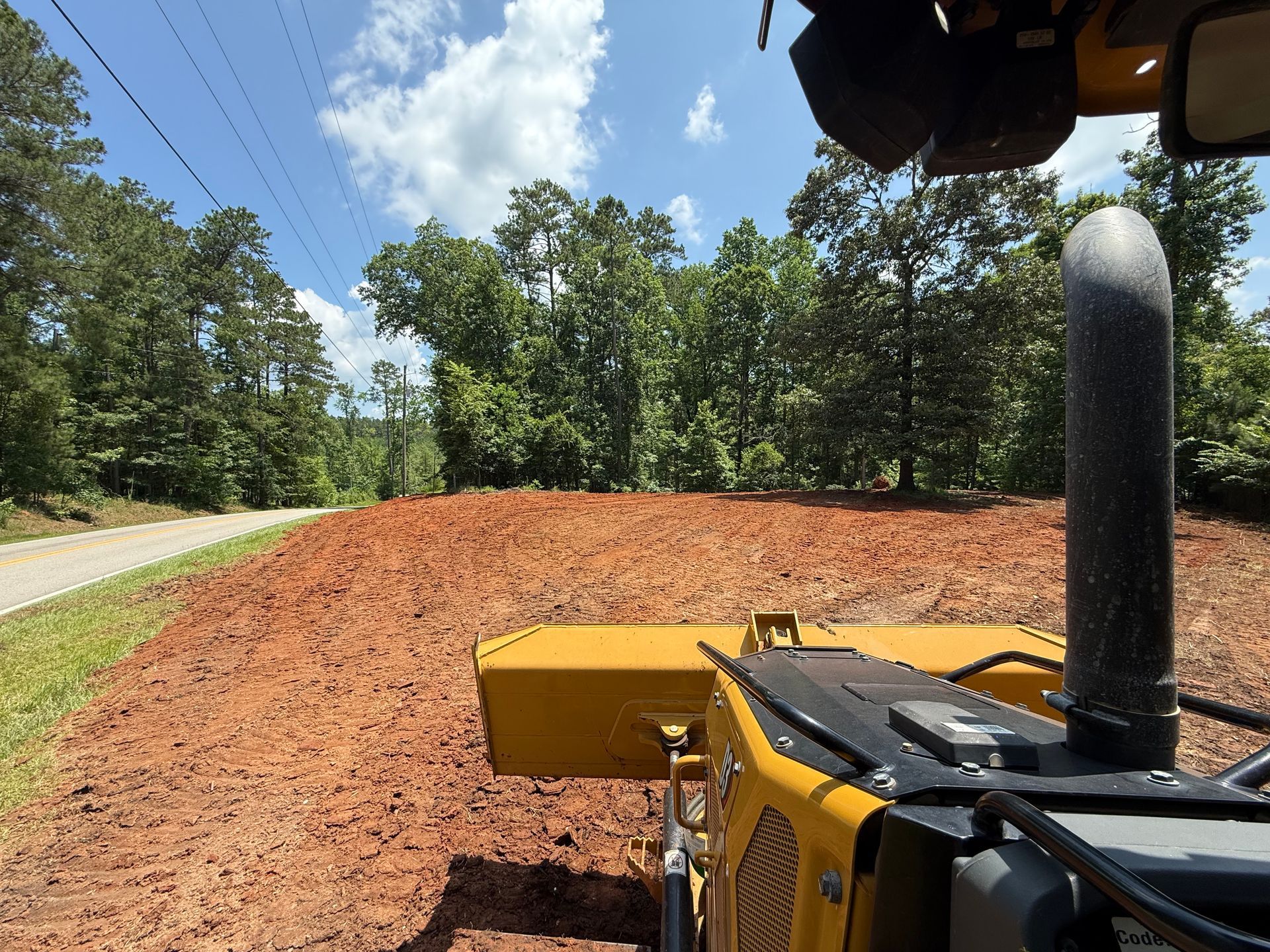 Yellow construction vehicle grading red dirt in a clearing next to a road, trees in the background.