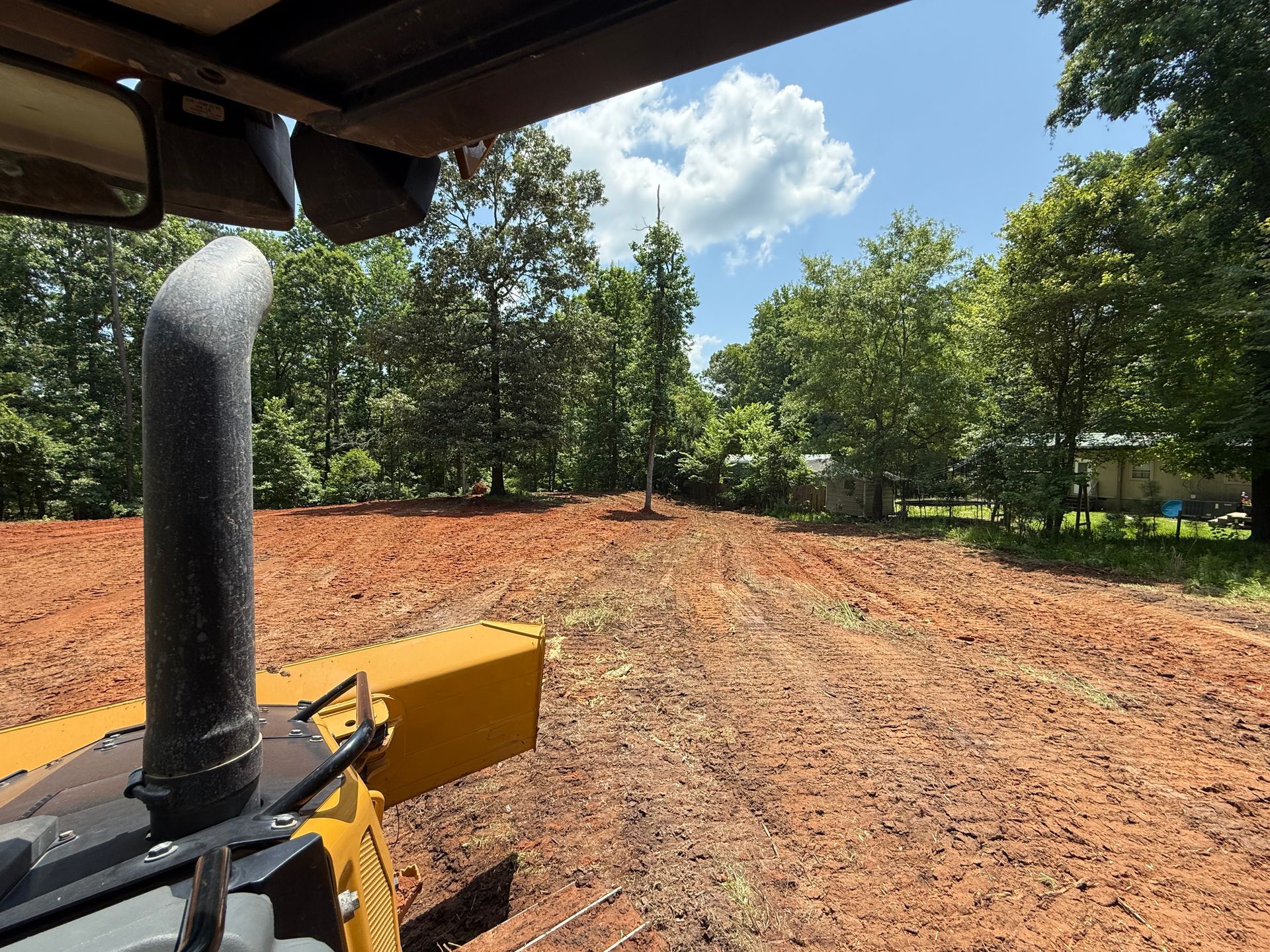 View from inside heavy machinery; a cleared, red dirt field with trees in the distance under a sunny sky.