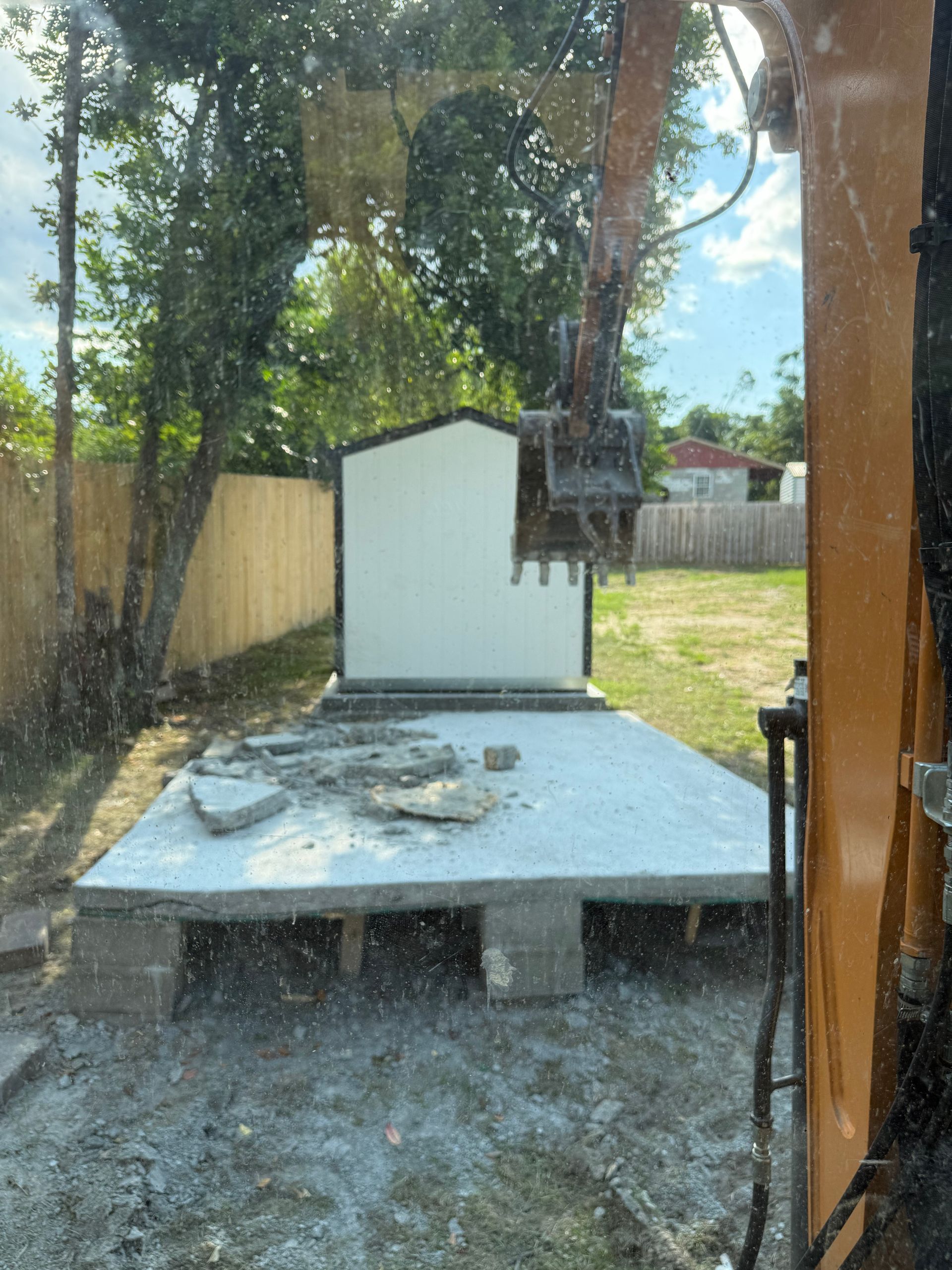 Excavator demolishes shed on concrete pad in yard; trees and fence in the background.