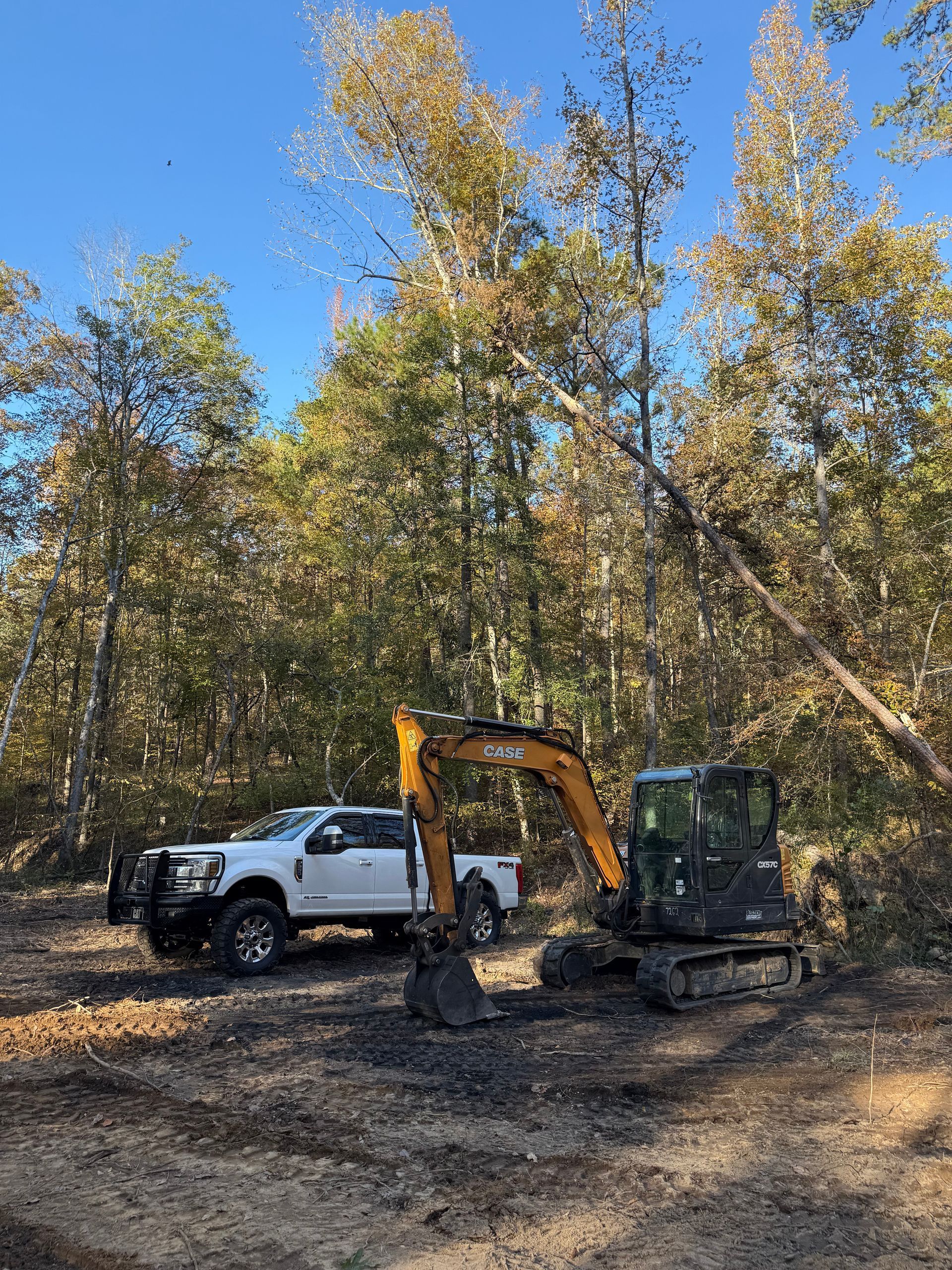 White truck and excavator in a clearing, trees with fall foliage in the background.