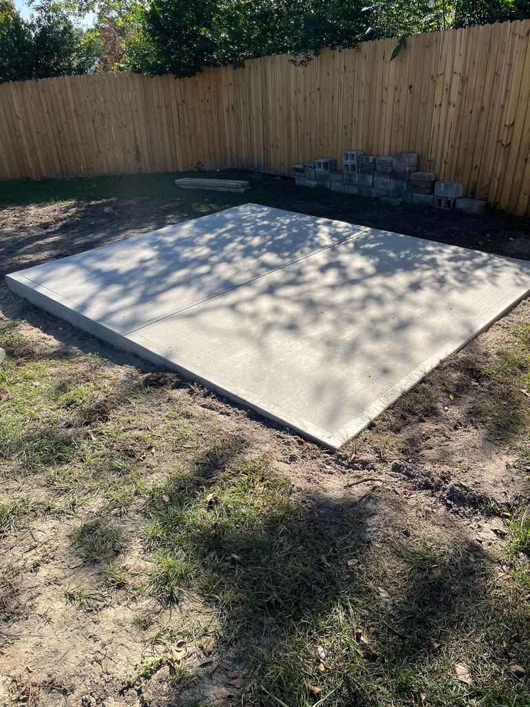 Square concrete slab in a grassy yard, near a wooden fence and small stone pile.