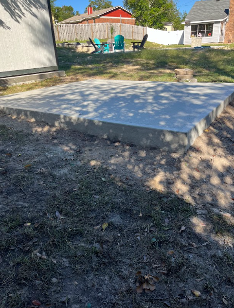 A rectangular concrete slab in a yard, surrounded by dirt and grass. A shed and chairs are in the background.