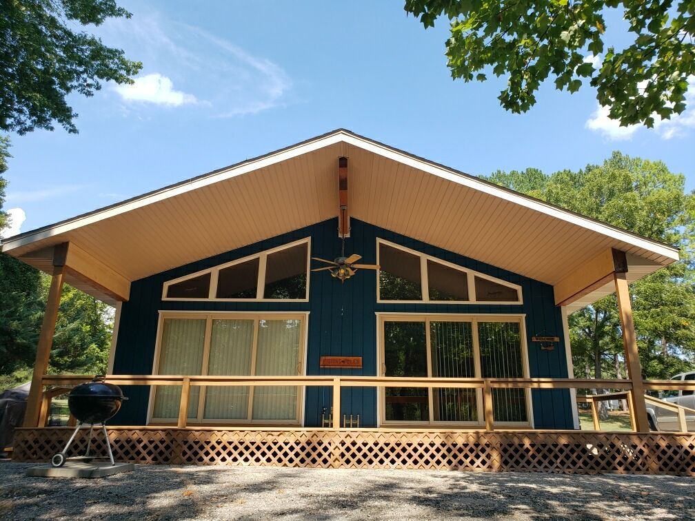 Blue cabin with a deck and large windows, surrounded by trees under a blue sky.