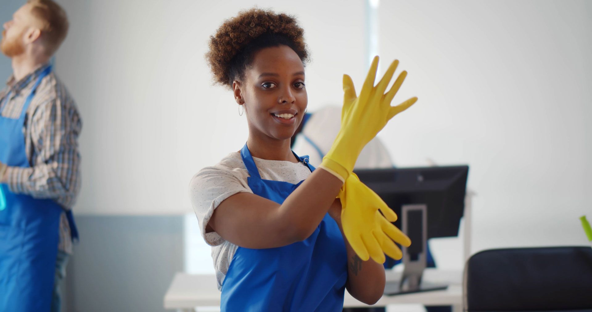 Woman in blue apron putting on yellow cleaning gloves, smiling.  Another person cleaning in background. Office setting.