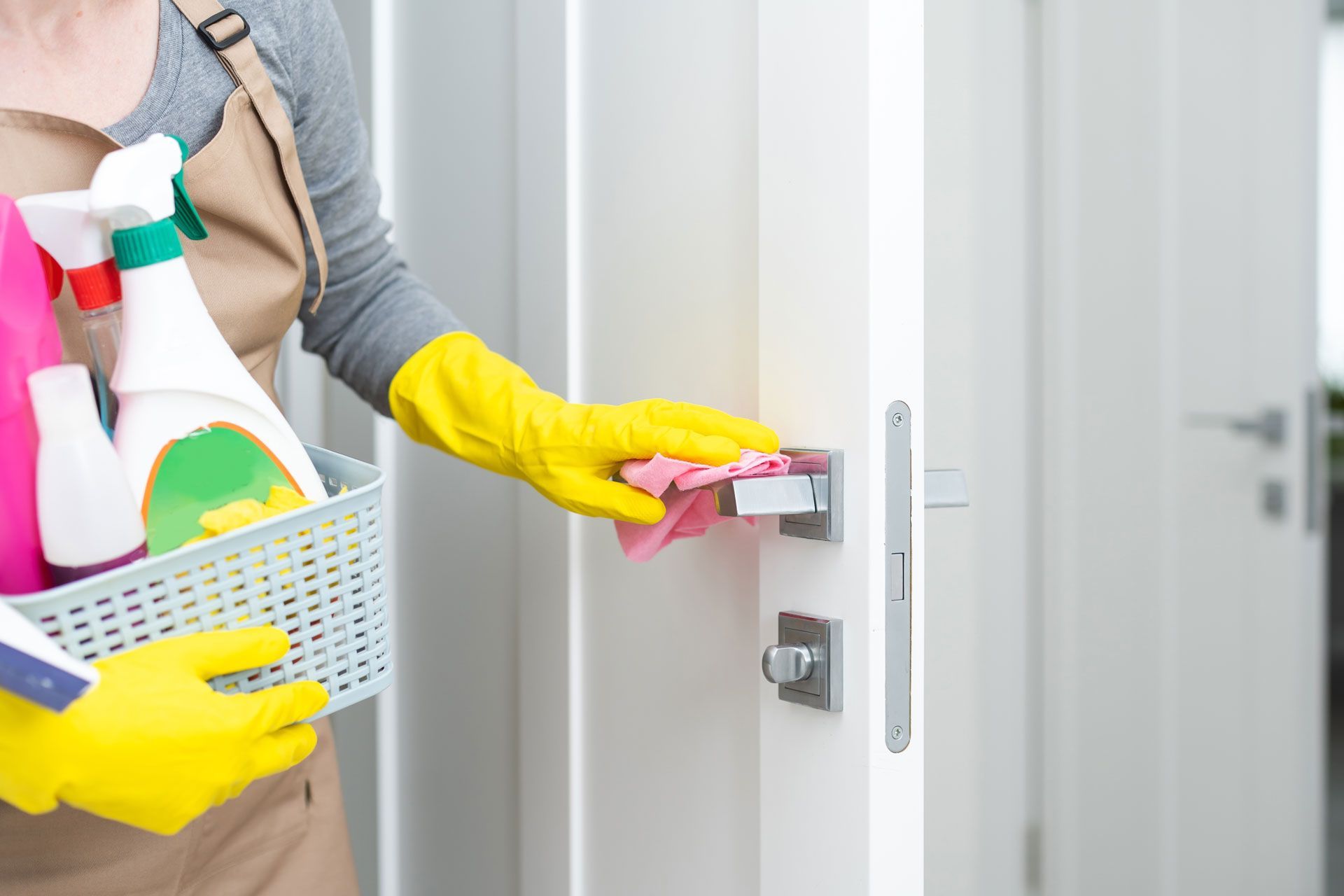 Person wearing yellow gloves wiping a door handle with a pink cloth, holding cleaning supplies in a basket.