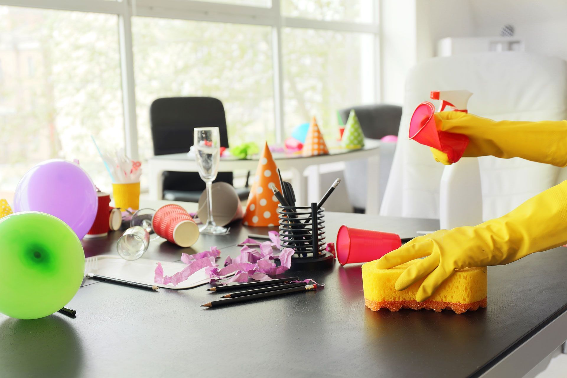 A person with yellow gloves cleans a messy office desk after a party, removing debris like cups and confetti.