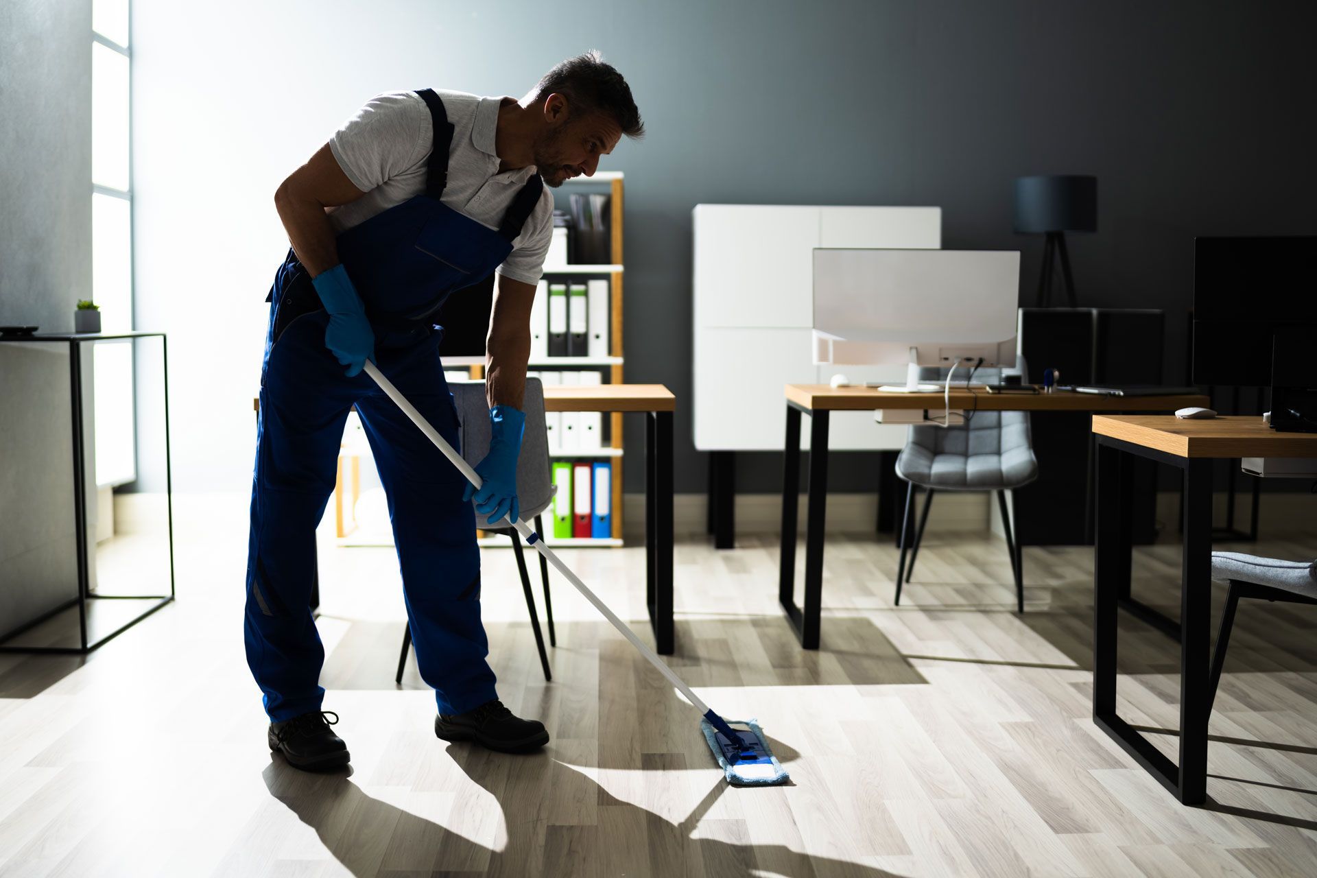 Man in blue cleaning overalls mopping an office floor with natural light.