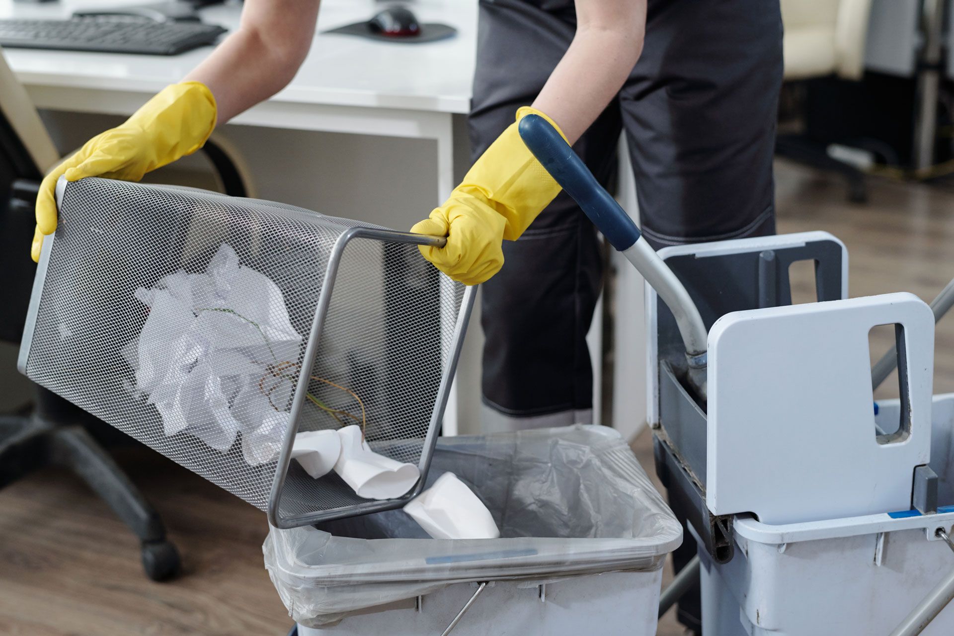 Person wearing yellow gloves emptying a metal wastebasket into a trash can on a cleaning cart in an office.