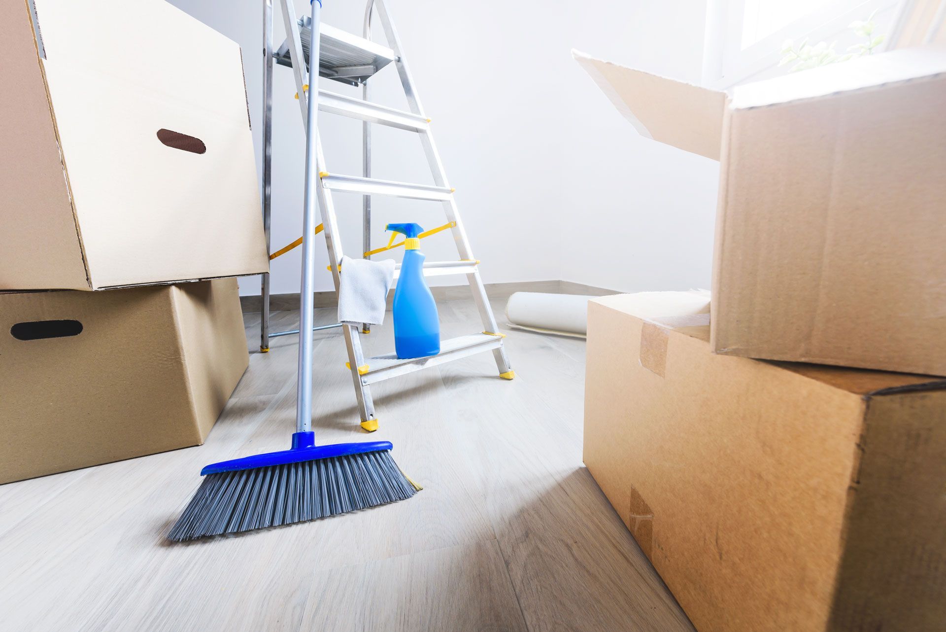 Moving boxes and cleaning supplies next to a ladder in a room with hardwood floors, suggesting a home move or renovation.