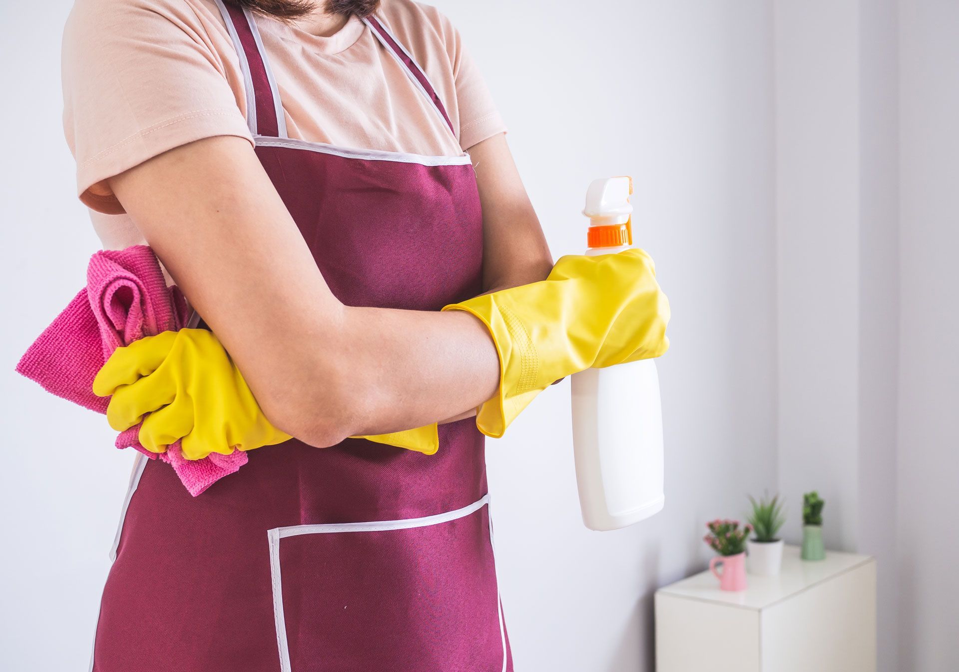 Person wearing an apron and yellow gloves holding a spray bottle and a pink cloth, ready to clean.