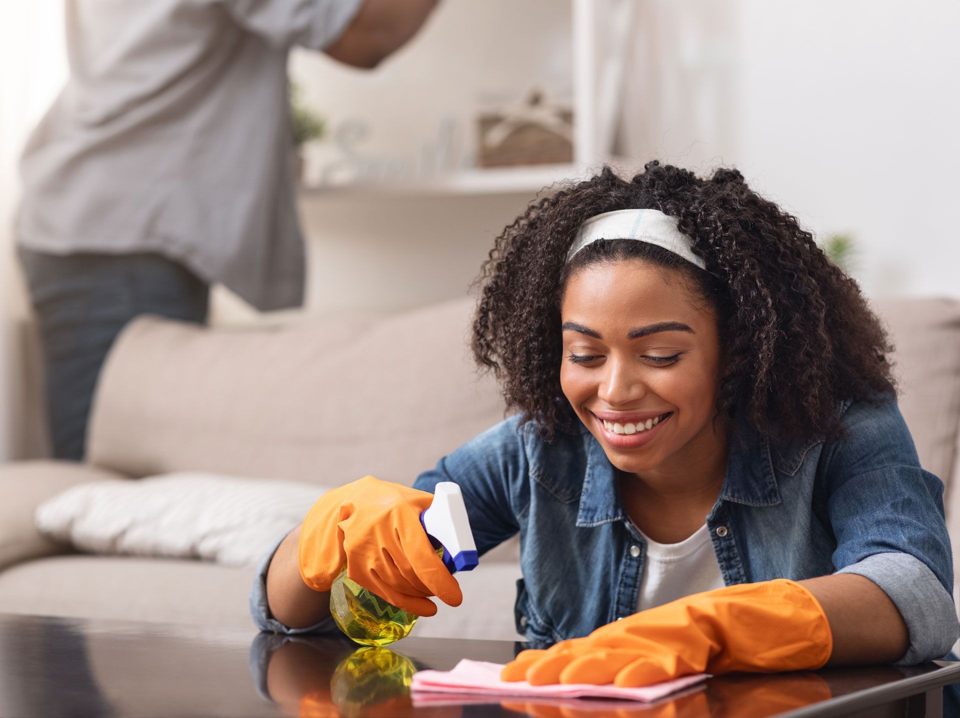 Woman wearing gloves, smiles while cleaning a table with spray bottle; man in background cleans shelves.