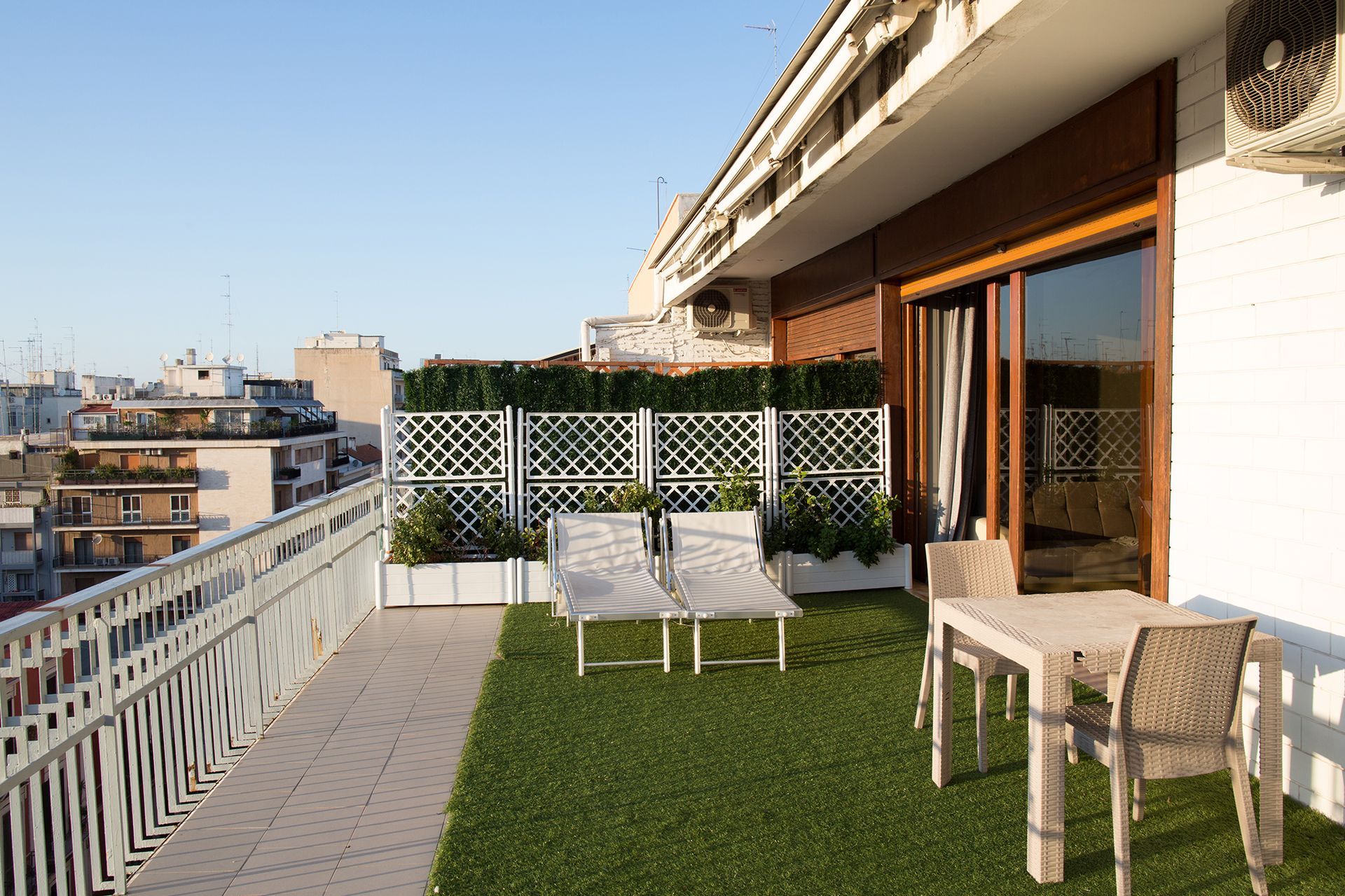 A sunny rooftop balcony with lounge chairs, a table, and cityscape views. The area has faux grass and a decorative trellis.