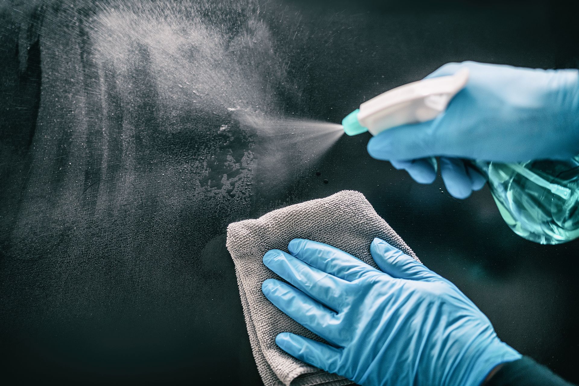 Hands in blue gloves spraying cleaner onto a surface and wiping with a gray cloth.