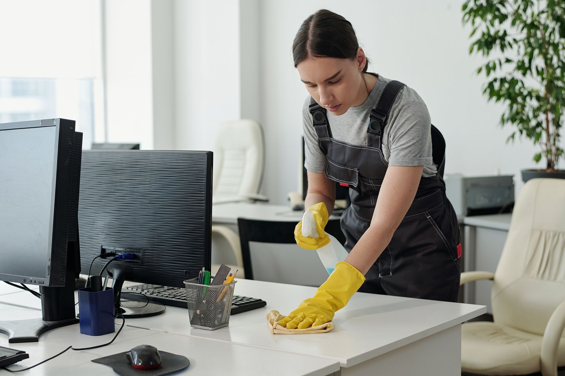 Woman in yellow gloves and overalls cleaning a white office desk with a spray bottle and cloth.