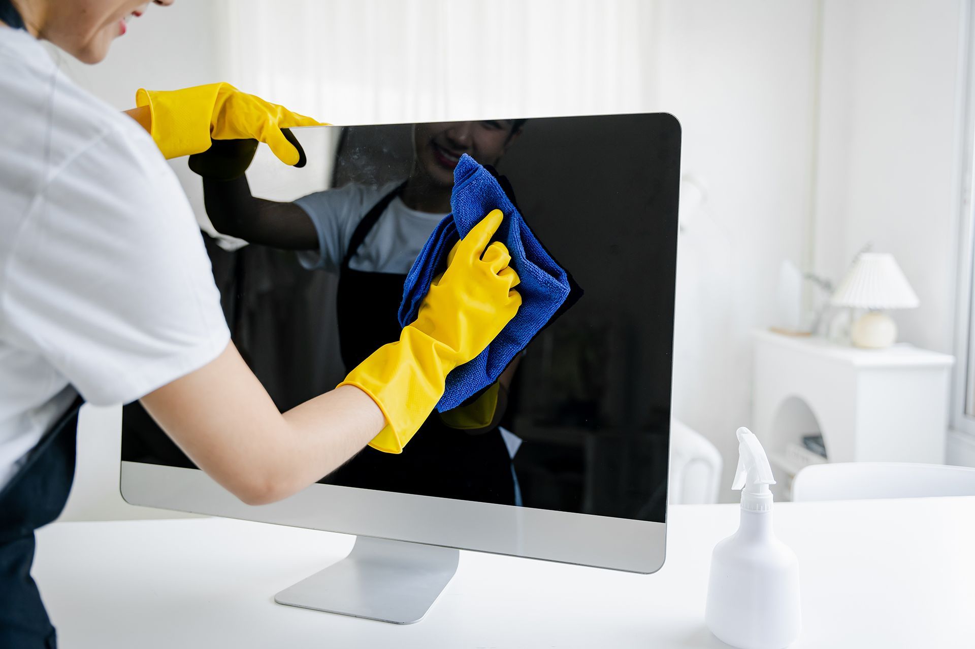 Person wearing yellow gloves cleaning a computer monitor with a blue cloth in a bright room.