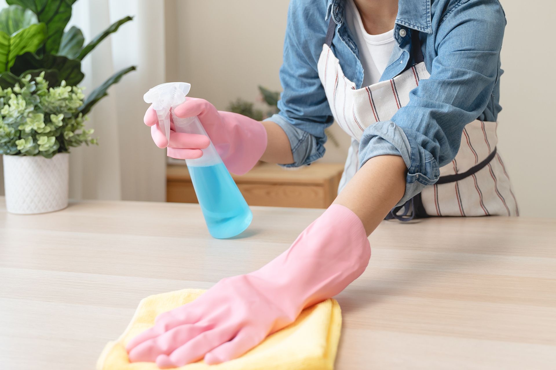 Woman wearing pink gloves and apron, spraying and wiping a wooden table with a yellow cloth.