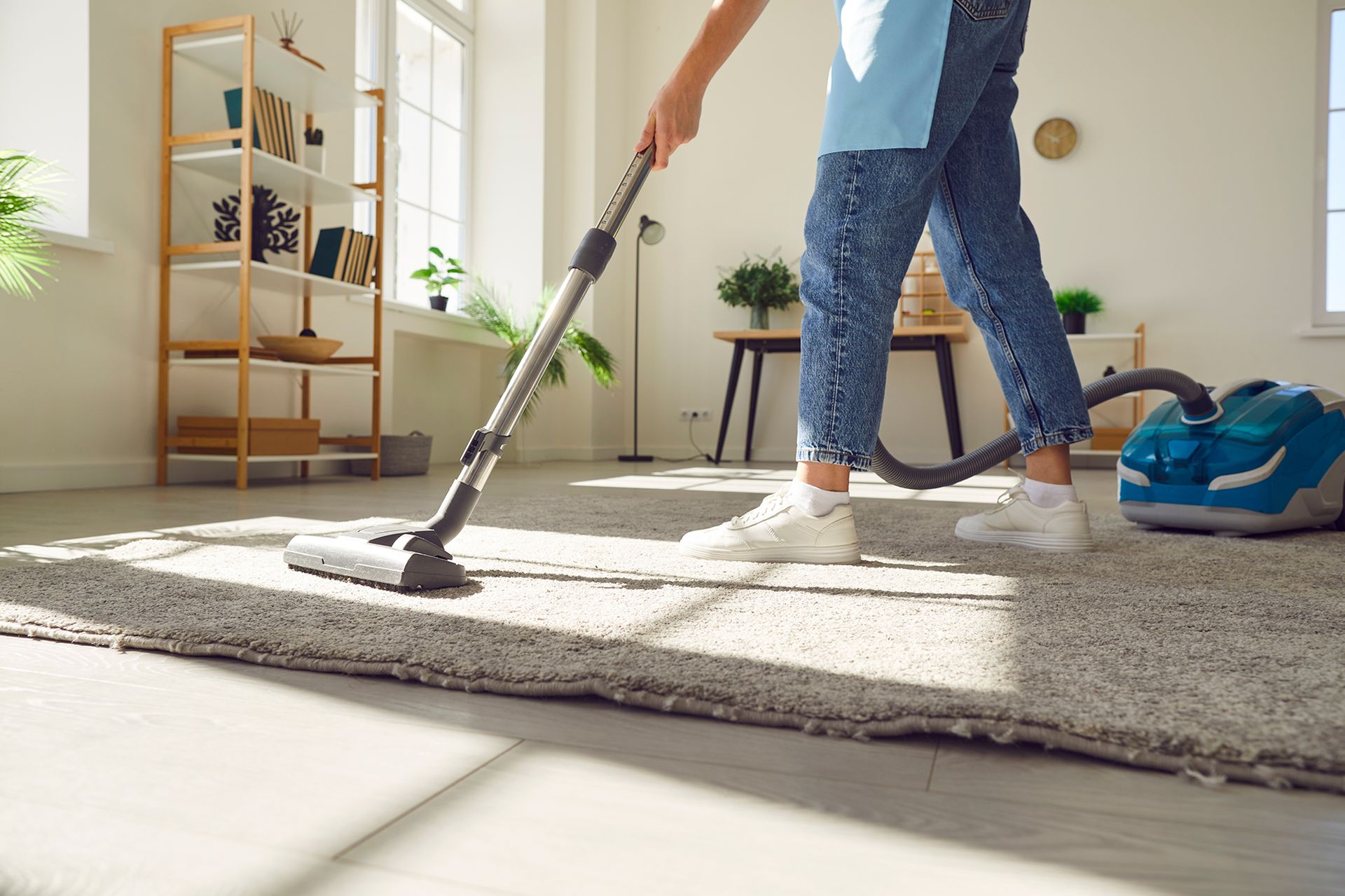 Person vacuuming a light-colored rug in a sunlit room. They wear jeans and white sneakers. A blue vacuum sits nearby.