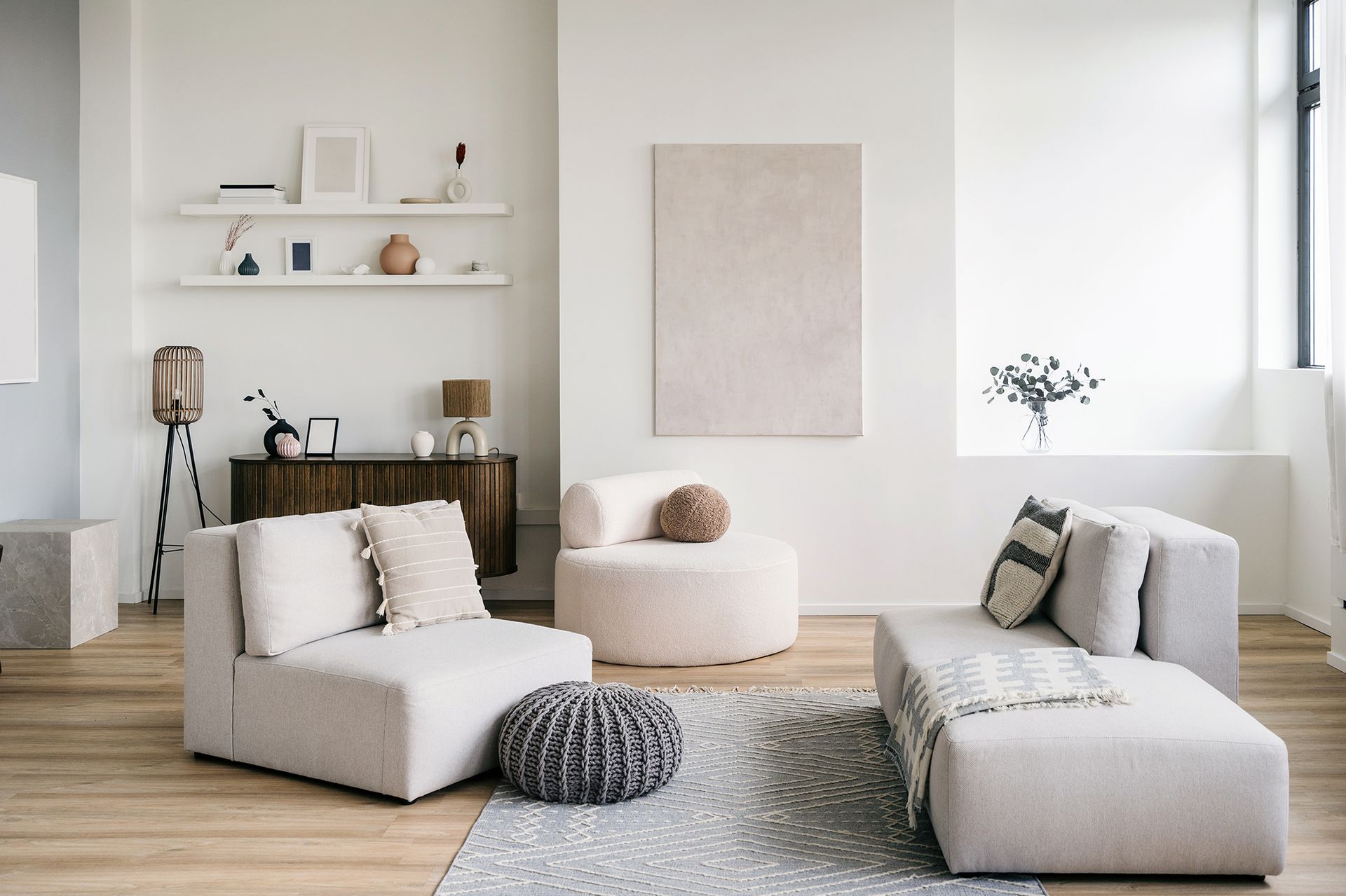 A modern living room with white walls, light-colored furniture, and decorative items like shelves and a large artwork.