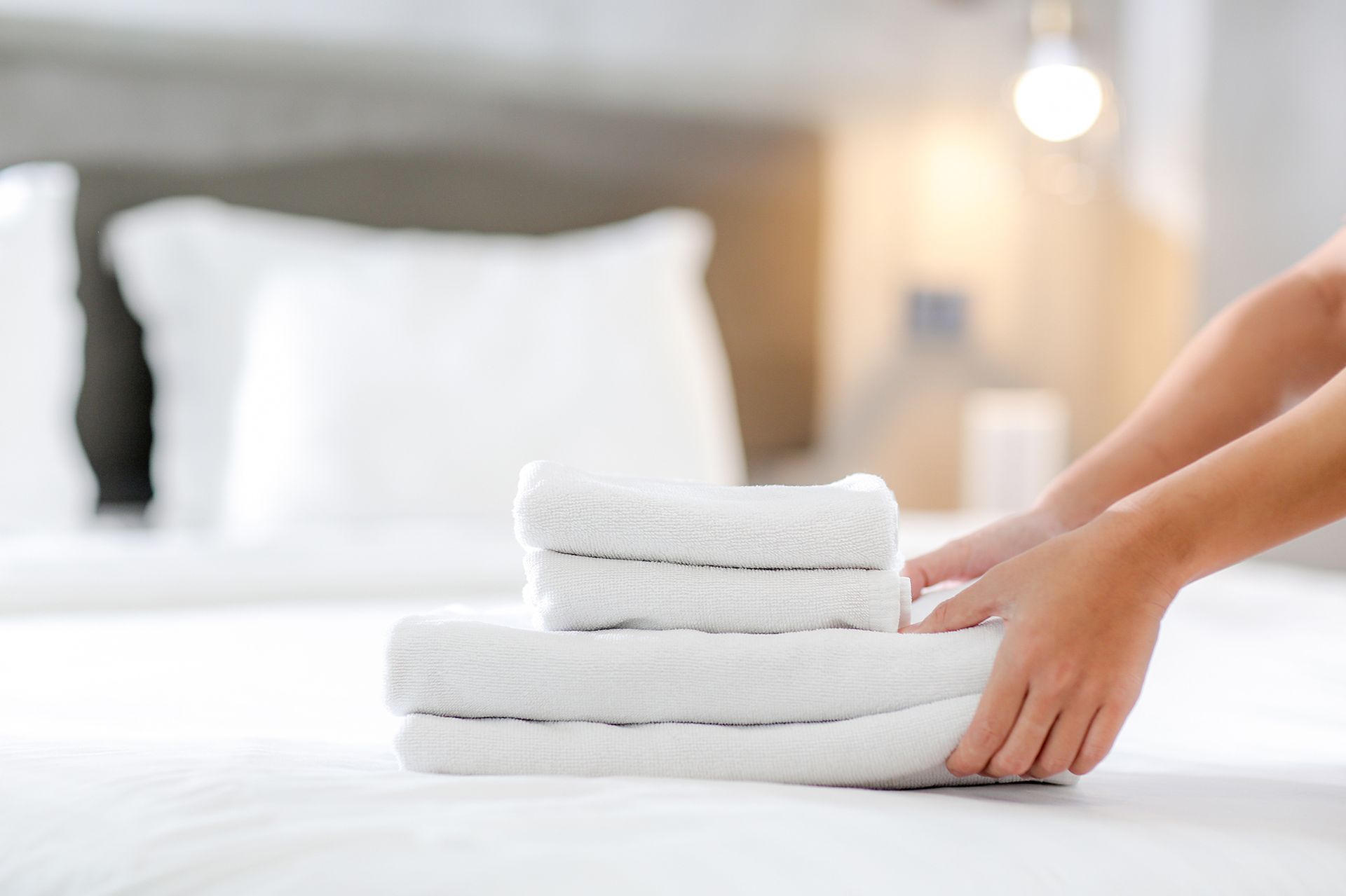 Person arranging a stack of white folded towels on a bed with a white comforter. Soft light and blurred background.