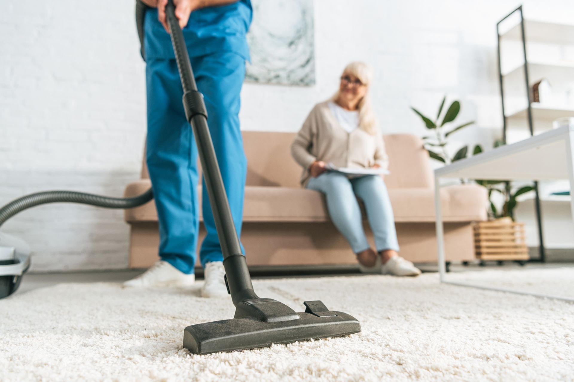 Person vacuuming a white carpet while a smiling older woman sits on a couch in a living room.
