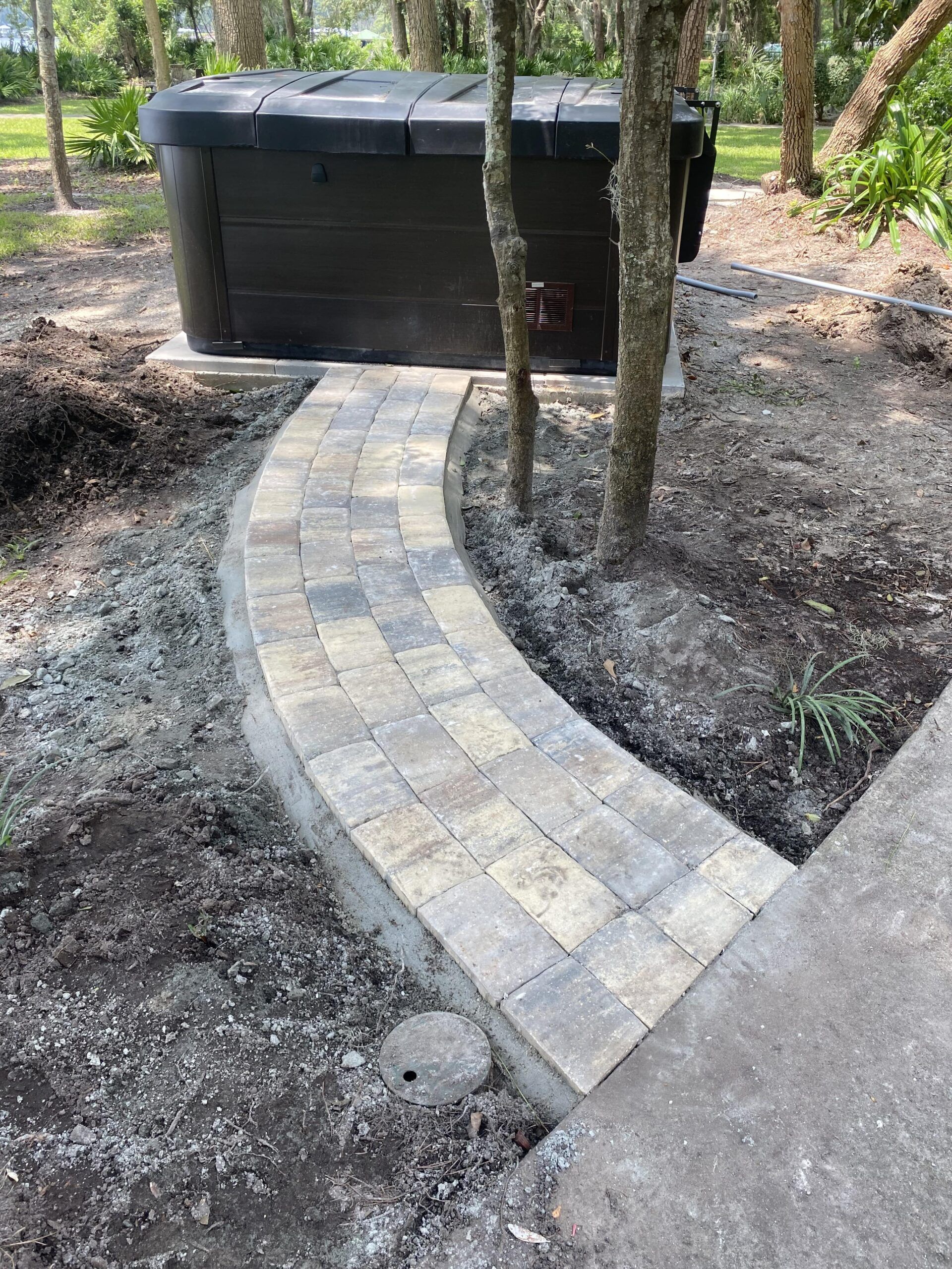 A brick walkway leading to a black trash can in the woods.