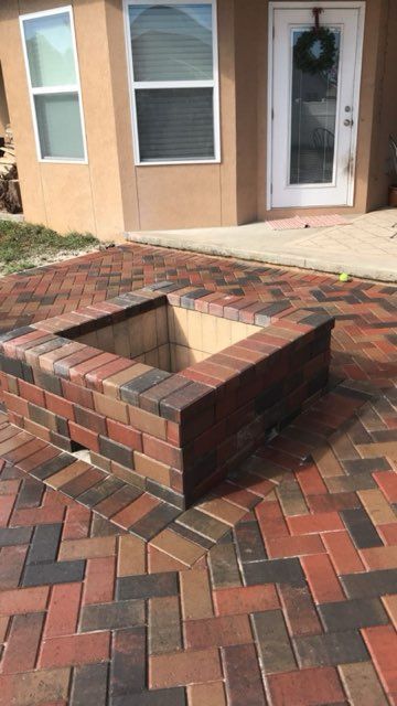 A brick fire pit is sitting on a brick patio in front of a house.