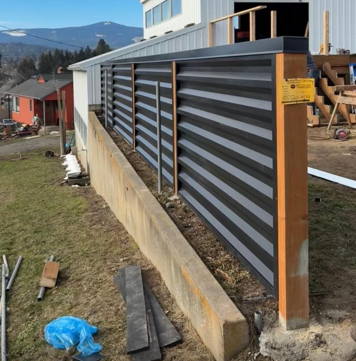 Dark gray horizontal corrugated fence along a concrete wall, next to a building and grass.
