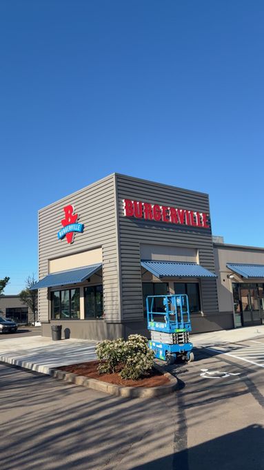 A person on a lift installs a sign on the Burgerville restaurant. Blue lift, red and gray building, clear sky.