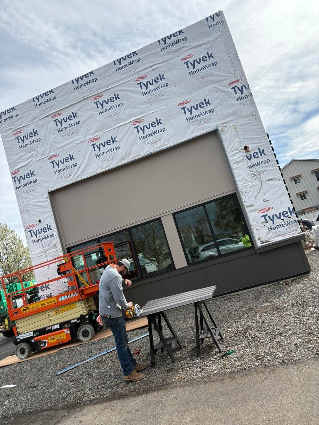 Building under construction. A worker stands near a table, next to a lift. The building is wrapped in Tyvek.