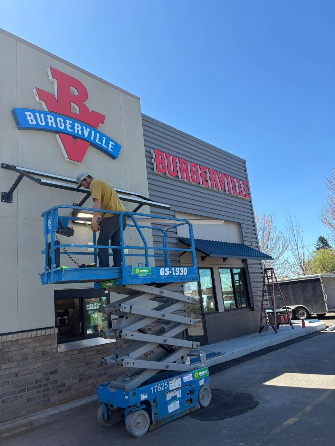 A person on a lift installs a sign on the Burgerville restaurant. Blue lift, red and gray building, clear sky.