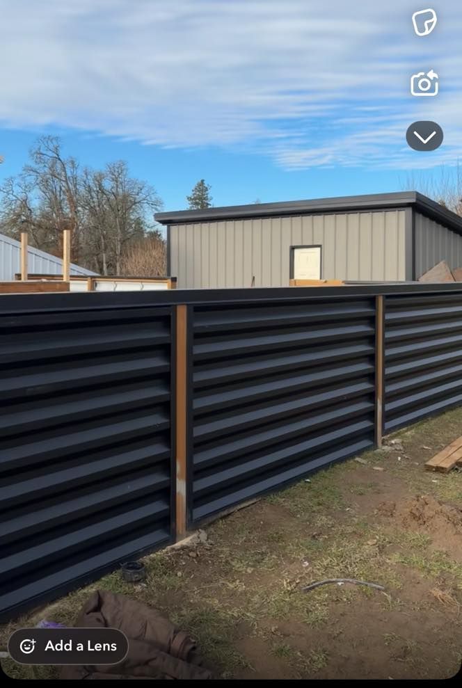 Black horizontal slatted fence with brown posts, set against a building and blue sky.