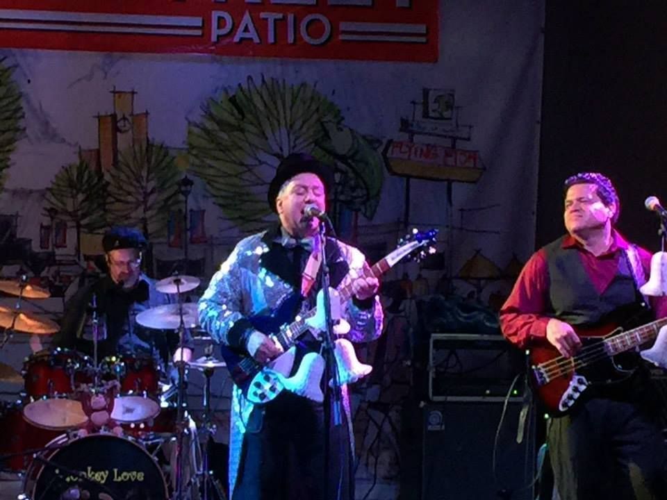 Band performing on a patio stage. A man in a silver jacket plays guitar and sings. A drummer and bassist accompany him.