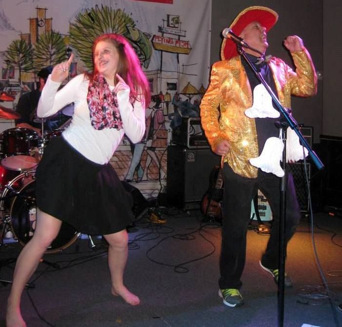 A woman and a man on stage, performing. The woman points, wearing a white top and skirt. The man in a gold jacket and cowboy hat gestures with microphone stand.