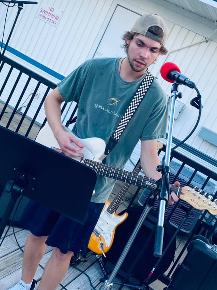 Man playing an electric guitar on an outdoor stage with a microphone. He wears a green shirt and backwards baseball cap.