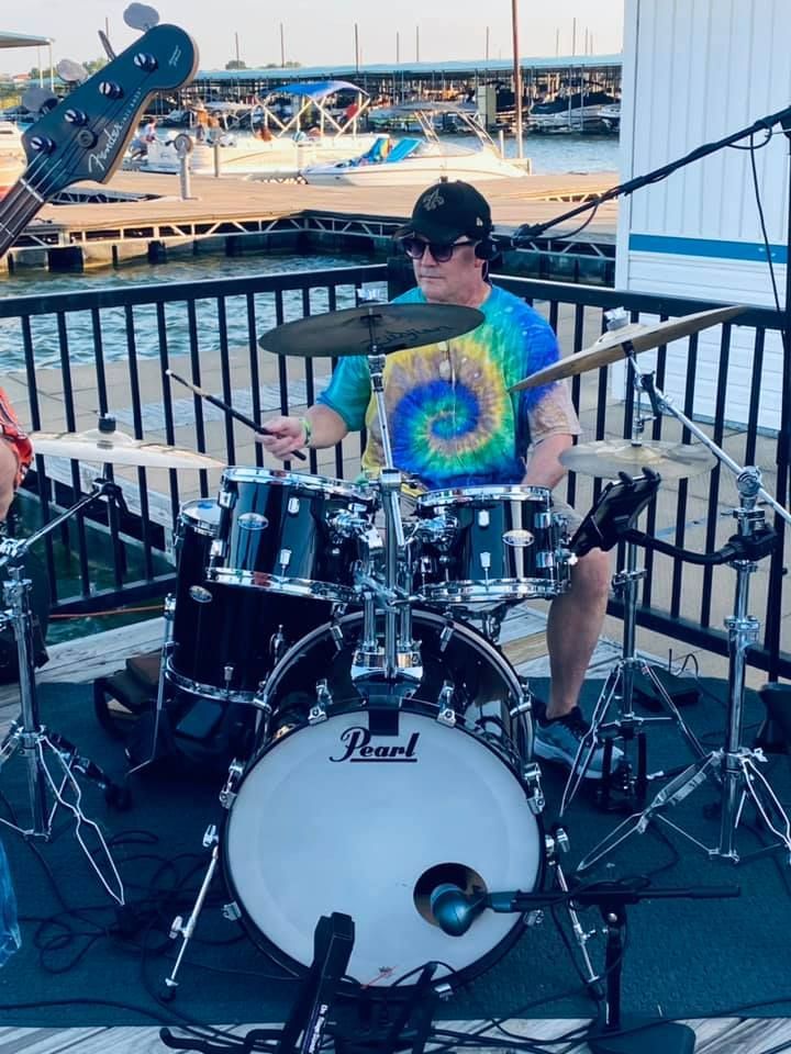Drummer playing outdoors on a black drum set. He's wearing a tie-dye shirt and sunglasses, with a waterfront setting in the background.