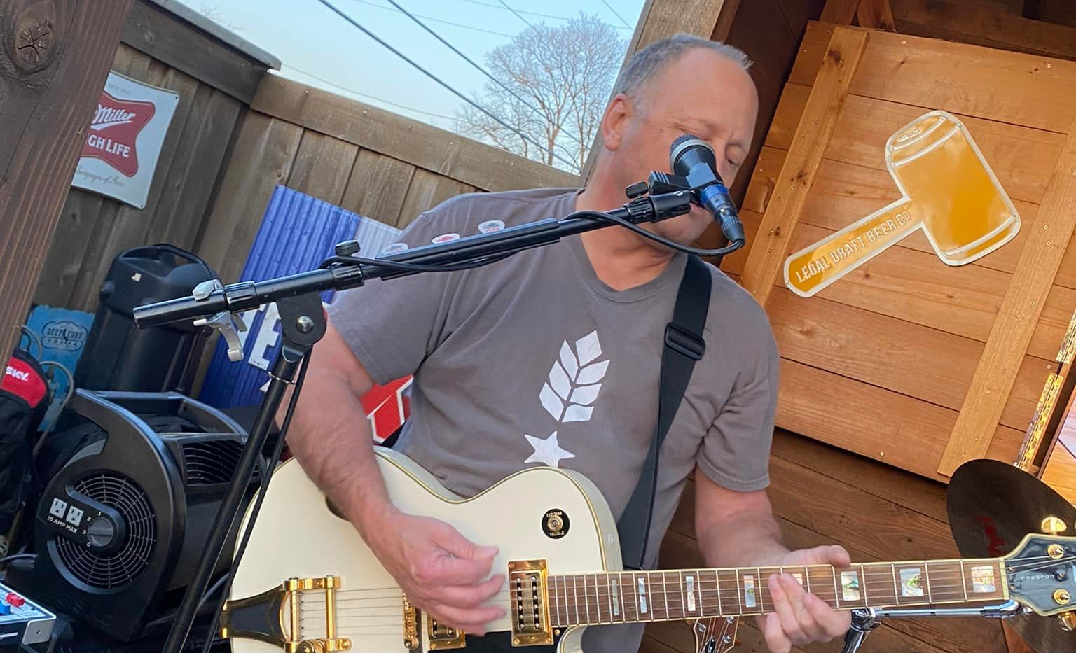 An outdoor restaurant patio at dusk. People sit at picnic tables under umbrellas strung with lights, near a large tree and a wooden fence.