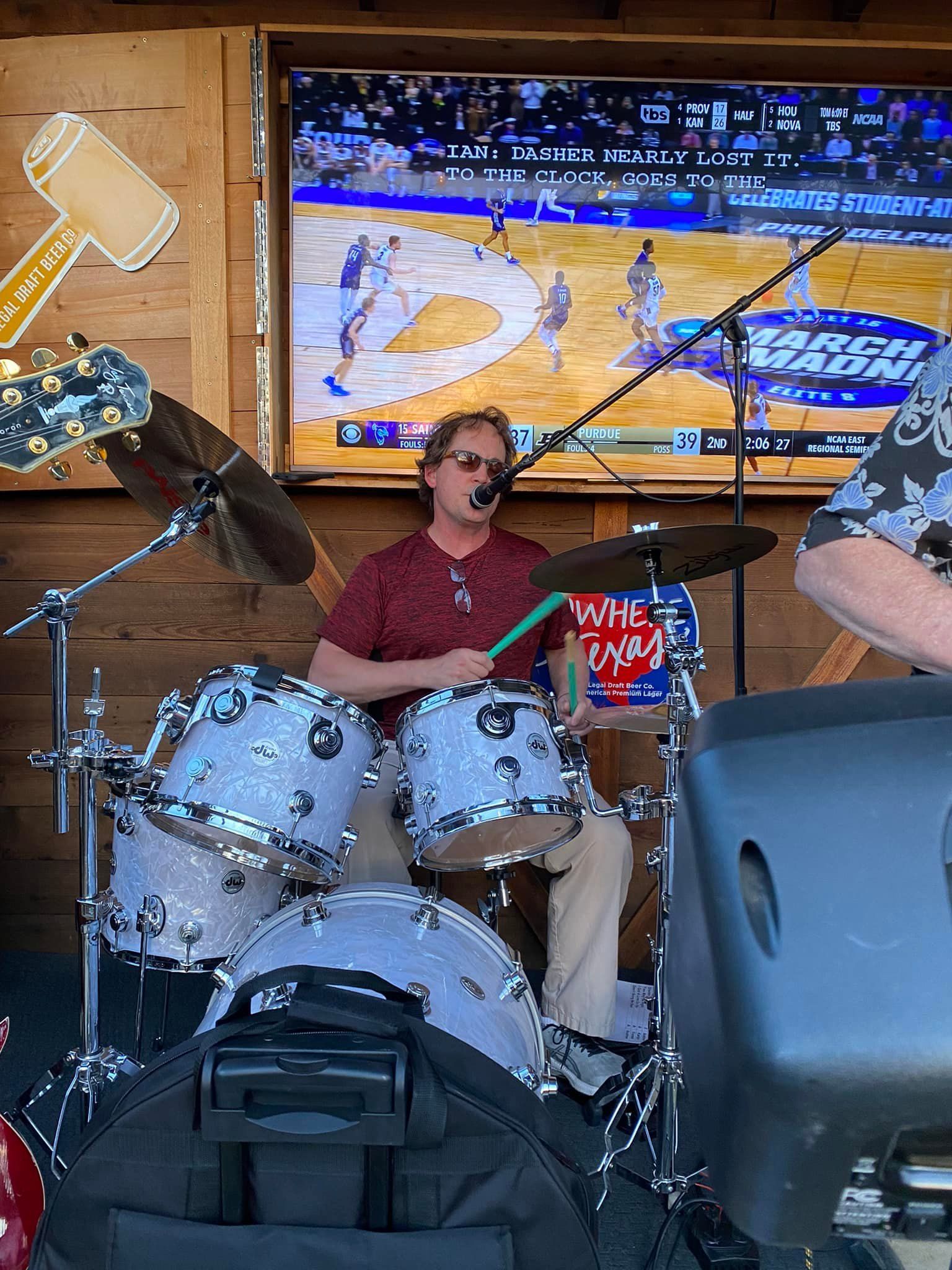 A drummer performs at an outdoor venue with a TV screen showing a basketball game. The drummer has light colored drums and wears a red shirt.