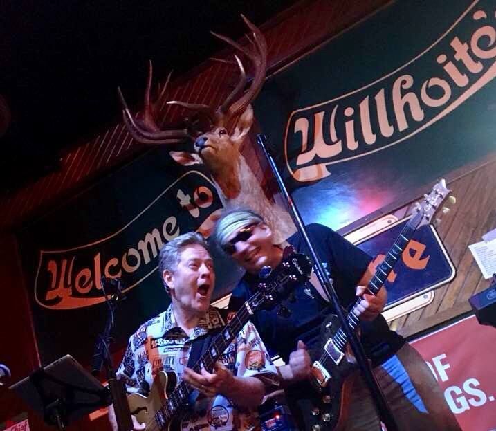 Two musicians performing on stage at Willhoite's bar. One sings with open mouth, the other plays guitar. A mounted deer head is above.