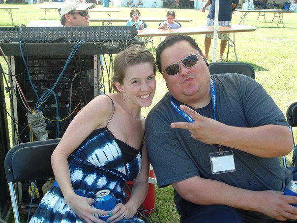 Woman and man smiling, posing together outdoors. Man makes peace sign, wearing sunglasses. Both hold cans, sitting near sound equipment.