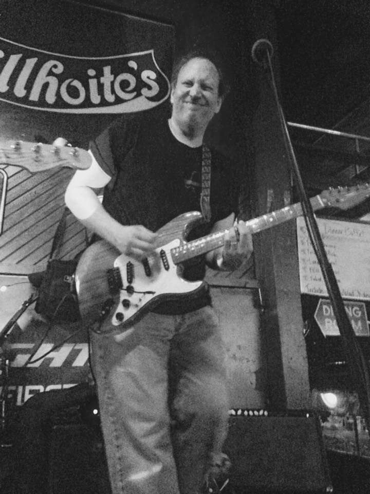 A smiling man playing electric guitar onstage at a bar. He wears a black shirt and jeans.