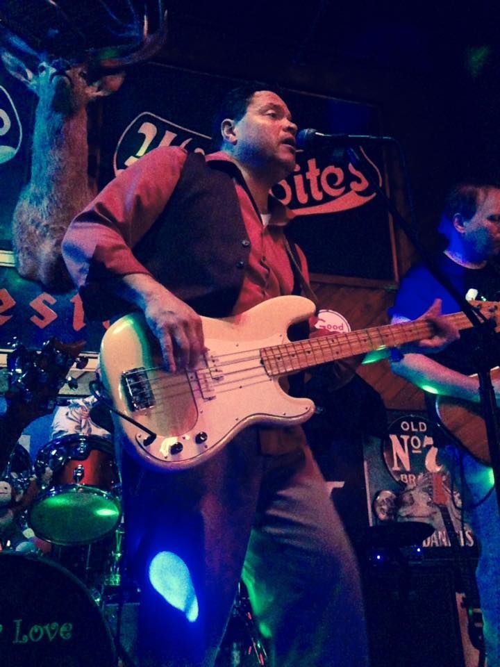 Man playing a white bass guitar, singing on stage at a bar. He wears a red shirt, black vest, and blue jeans.