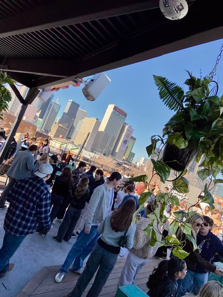 People gather on an outdoor rooftop with a city skyline in the background on a sunny day. Hanging plants and overhead beams are visible.