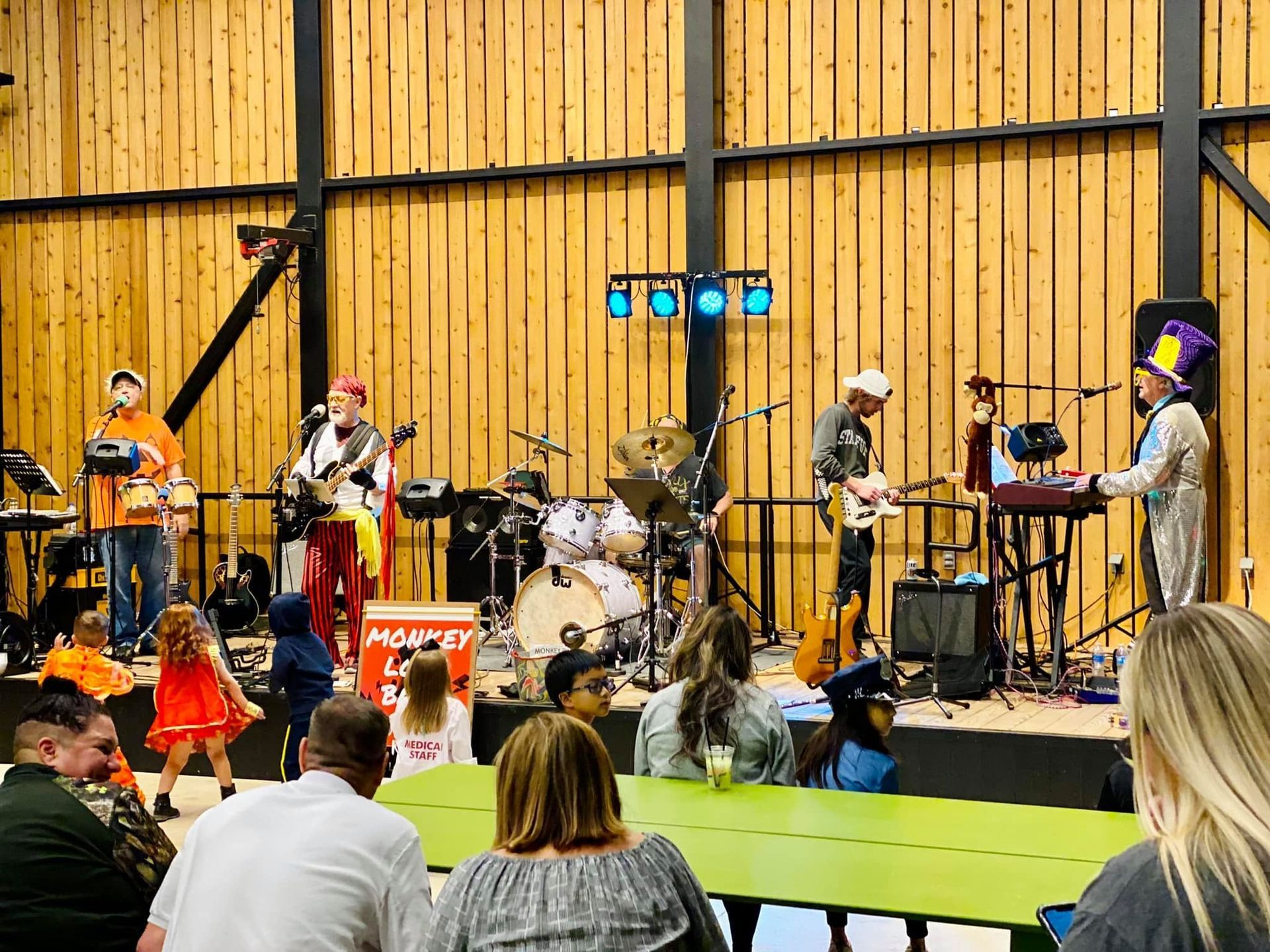A band performs on stage at an indoor event. Audience members watch from tables. The stage is brightly lit with colorful costumes.