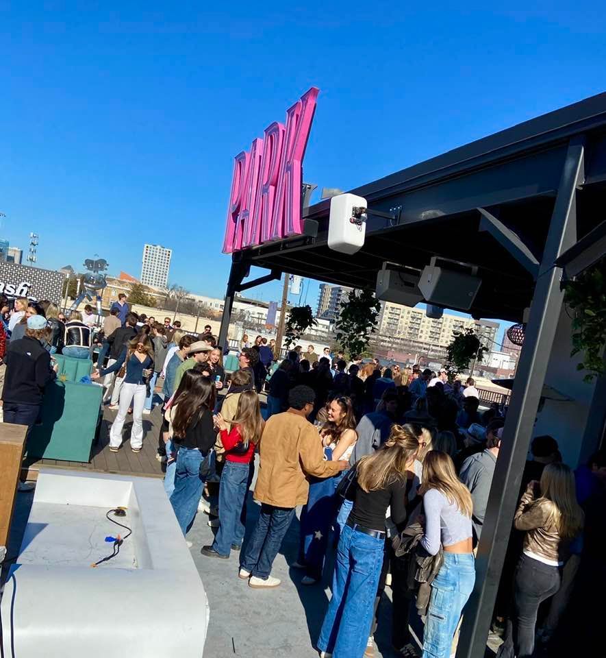 People crowd outside a bar with a pink sign that reads 