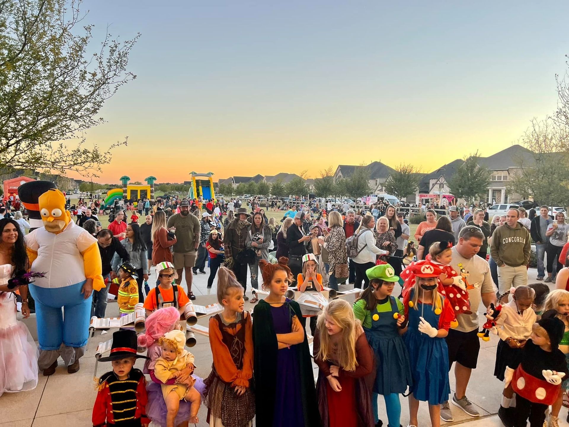 Halloween gathering outdoors at dusk. Children in costumes stand in a line; adults and other children mingle nearby with bouncy castles in the distance.